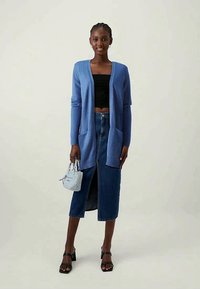 Blå strikket cardigan med lange ermer, åpen front og to fremre lommer, kombinert med en svart crop top og blå denimskjørt. Sorte sandaletter med hæl og en liten blå veske.