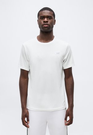 Giovane uomo nero che indossa una maglietta bianca a maniche corte semplice e pantaloni bianchi, in piedi davanti a uno sfondo grigio chiaro neutro.