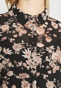 Czarna bluzka w kwiaty z kołnierzykiem, z zapięciem na guziki i jasnoróżowymi wzorami kwiatowymi na przezroczystym materiale.