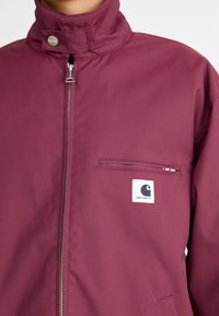 Bordeauxkleurige jack van duurzaam materiaal, met een hoge kraag, een ritssluiting aan de voorkant en een borstzak voorzien van een logo-embleem.