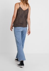 Kobieta w brązowej bezrękawnikowej bluzce w leopardzi wzór, jasnoniebieskich dżinsach z surowymi brzegami i czarnych wysokich trampkach, odchodząca na białym tle.