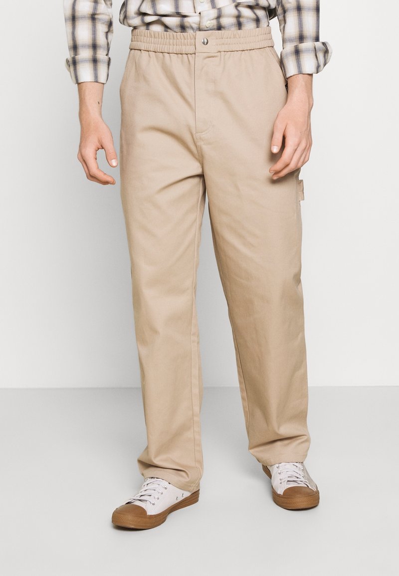Pantalon en coton beige avec une taille élastique, des jambes droites et une poche latérale, porté avec des baskets blanches et une chemise à carreaux.