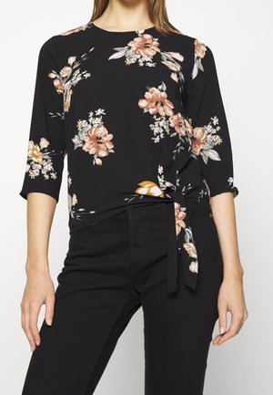 Femme portant un haut noir à imprimé floral avec des fleurs roses et blanches, manches trois-quarts, et un nœud noué à la taille, associé à un pantalon noir.