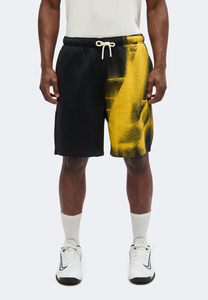 Homme portant un short noir et jaune à motifs avec un cordon blanc, des chaussettes blanches et des chaussures de sport blanches, debout devant un fond uni.