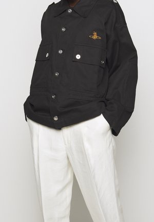 Person trägt eine schwarze, mit Knöpfen versehene Jacke mit Brusttaschen und einem gold bestickten Logo, kombiniert mit lockeren weißen Hosen, Hände in den Taschen.