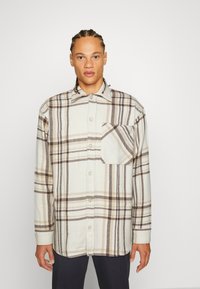 Camicia di flanella chiara con un motivo a quadri in marrone e crema, con chiusura a bottoni davanti e taschino sul petto. Maniche lunghe.