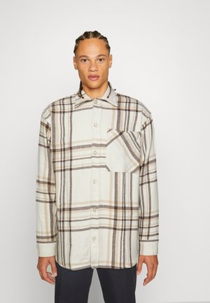 Camicia di flanella chiara con un motivo a quadri in marrone e crema, con chiusura a bottoni davanti e taschino sul petto. Maniche lunghe.