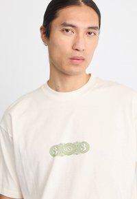 Persona con cabello oscuro recogido que lleva una camiseta blanca con un diseño ornamentado en verde que presenta el número "3", la palabra "UNO" y otro "3" en el centro.