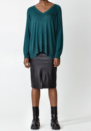 Femme portant un pull ample à col en V couleur sarcelle, une jupe en cuir noire mi-genou et des bottines noires épaisses, debout devant un fond blanc.