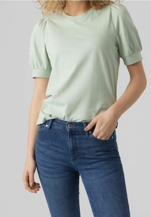 Femme portant un chemisier vert clair à manches courtes et bouffantes et un jean bleu, une main dans la poche, sur fond uni.