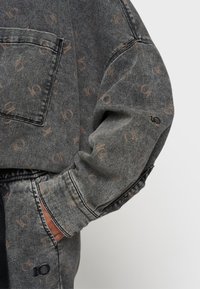 Giacca di jeans in grigio con un sottile motivo a logo marrone. Presenta una tasca frontale e una vestibilità rilassata con maniche a polsino. Tessuto testurizzato.