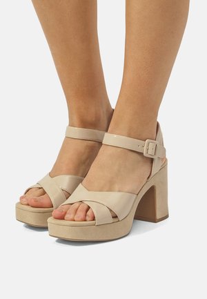 Pies con sandalias de plataforma beige con tacones gruesos, correas en el tobillo y tiras cruzadas en la parte delantera sobre un fondo blanco.