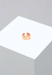 Anello in rame con una finitura lucida e un'apertura intagliata, adagiato su un pedestal bianco luminoso su uno sfondo minimalista.