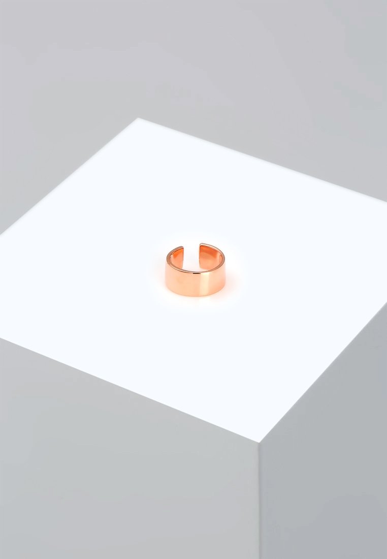 Anello in rame con una finitura lucida e un'apertura intagliata, adagiato su un pedestal bianco luminoso su uno sfondo minimalista.