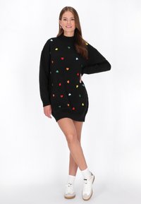 Schwarzes Strickkleid mit hohem Kragen, das durchgehend mit bunten Herzmustern gestaltet ist. Kombiniert mit weißen Sneakers und Socken.