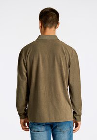 Camicia a coste verde oliva con maniche lunghe e colletto, caratterizzata da un tessuto texturizzato e un orlo dritto, indossata sopra jeans blu.