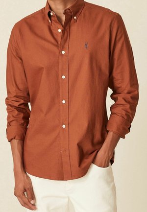Mężczyzna ubrany w rdzawy koszulę na guziki z białymi guzikami i małym haftowanym logo, w połączeniu z białymi spodniami, ręką w kieszeni.