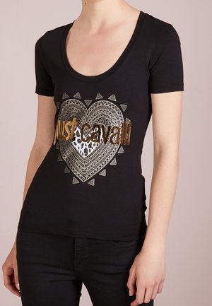 Vrouw draagt een aangesloten zwarte korte mouw shirt met een gouden en zilveren geometrisch hart en "Just Cavalli" tekst aan de voorkant.