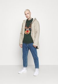 Beige jack met capuchon over een donkergroene gebreide trui met een rendierontwerp, blauwe jeans en witte sneakers. Effen achtergrond.