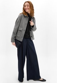 Grau wollene Jacke mit einem Kragen und einer Fronttasche, kombiniert mit einem schwarzen Oberteil, weiten navyblauen Hosen und schwarzen Ballerinas. Hält eine strukturierte navyblaue Clutch.