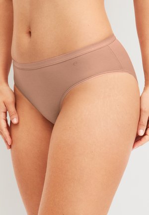 Femme portant une simple lingerie beige taille haute, debout les mains détendues le long du corps, fond neutre.