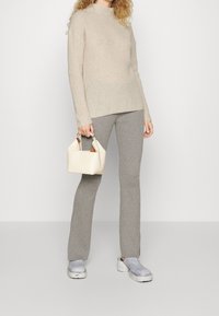 Femme portant un pull en tricot beige, un pantalon gris à côtes, des chaussures à plateforme blanches, tenant un sac à main de couleur crème sur un fond uni.
