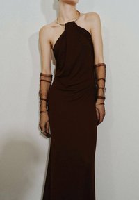 Brązowa sukienka na karku z materiałem w drapowania, przezroczyste brązowe rękawy oraz metalowy detal przy kołnierzu. Gładka tekstura, dopasowana sylwetka, długość do ziemi.