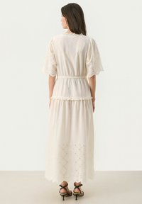 Maxi dress in light cream fabric with embroidered details, elastic waistband, and scalloped hem featuring diamond cutouts. Worn with black sandals.