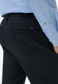 Schwarze, strukturierte Hose mit glatter Oberfläche, ausgestattet mit einer Gesäßtasche mit Knopfverschluss und einem kleinen Markenetikett darüber.