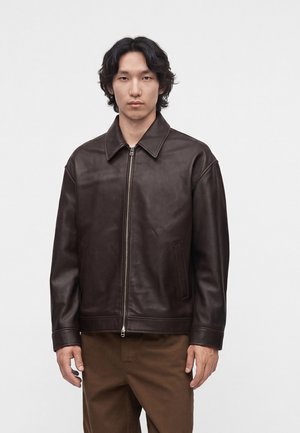 Jeune homme aux cheveux noirs mi-longs portant une veste en cuir marron foncé avec une fermeture éclair sur le devant et un pantalon marron sur un fond uni.