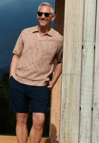 Polo à manches courtes en tricot texturé de couleur pêche clair, associé à un short bleu marine. Des lunettes de soleil portées, appuyé contre un mur en bois.