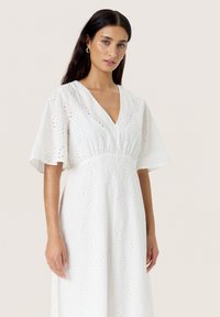 White dress featuring a V-neck, short wide sleeves, and an eyelet pattern with diamond shapes. Lightweight, textured fabric.
