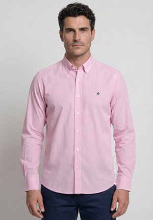 Hombre que lleva una camisa rosa claro con botones y pequeño logo oscuro en el pecho, pantalones azul marino, de pie frente a un fondo liso.