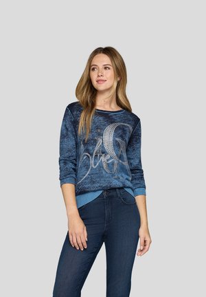 Blauer gestrickter Pullover mit grafischem Design und silbernen Akzenten. Gerippte Bündchen und Saum, strukturierter Stoff, kombiniert mit dunkelblauen Jeans.