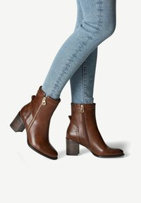 Bottines en cuir marron avec un talon carré, fermeture éclair sur le côté et clous décoratifs. Texture lisse avec un design élégant.