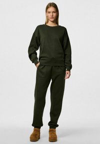 Felpa oversized verde scuro con polsini e orlo a costine, abbinata a pantaloni jogger coordinati con polsini elasticizzati e tasche laterali, indossata con scarpe con plateau marroni.