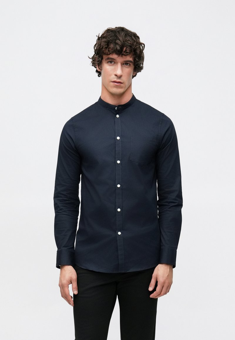 Μπλε ναυτικό πουκάμισο με κουμπιά, υψηλό γιακά, μακριά μανίκια και τσέπη στο στήθος· διαθέτει αντίθετα λευκά κουμπιά και λεία υφή υφάσματος.