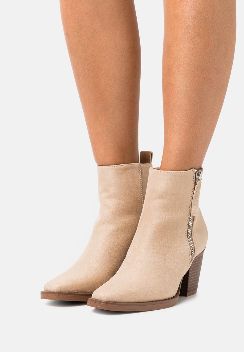 Bottines beiges en daim avec bouts pointus, fermetures éclair latérales et talons blocs en bois empilés, portées sur des jambes nues.