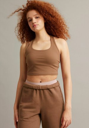 Młoda kobieta z kręconymi rudymi włosami, ubrana w brązowy krótki top i pasujące brązowe spodnie dresowe na jednolitym tle.