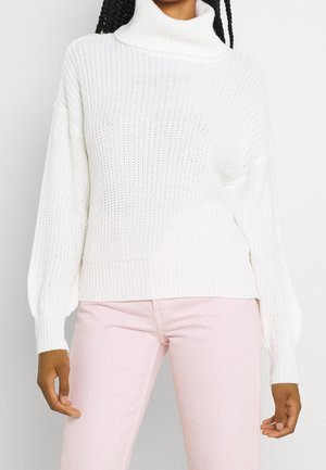 Femme portant un pull à col roulé en maille blanche et un pantalon rose clair, debout, les bras détendus le long du corps.