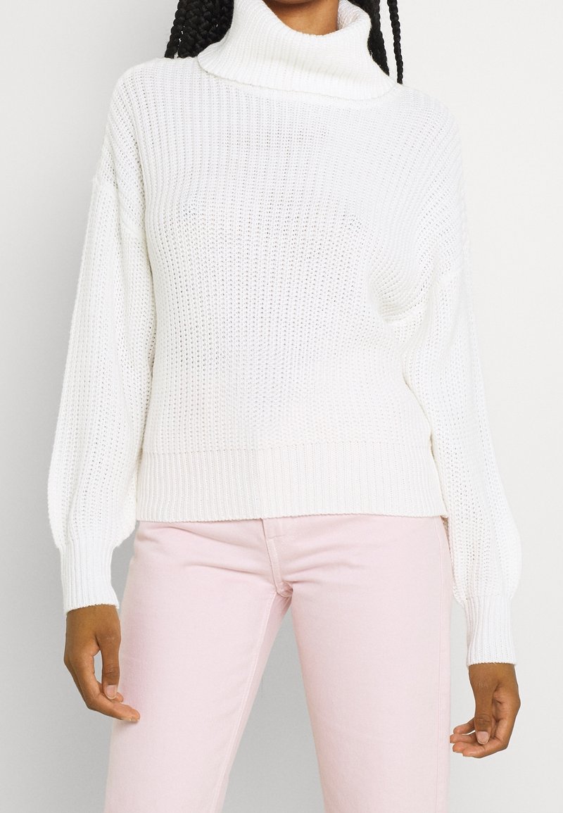 Femme portant un pull à col roulé en maille blanche et un pantalon rose clair, debout, les bras détendus le long du corps.