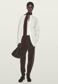 Chemise rayée blanche, écharpe bordeaux, pantalon large marron, tenant un sac à main marron foncé, portant des sandales noires. Silhouette simple et moderne.