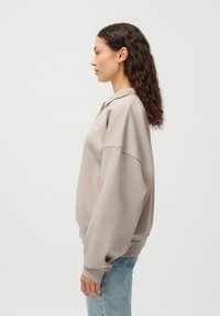 Lys beige oversized genser med en myk tekstur, med krage og subtil logo. Kombinert med lyseblå jeans for et uformelt utseende.