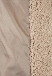 Zacht beige stof naast een gestructureerd beige sherpa fleece materiaal, die het contrast laat zien tussen gladde en pluizige oppervlakken.