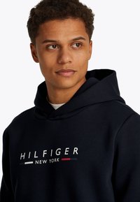 Jauns vīrietis ar īsiem cirtainiem matiem, valkājot tumši zilu kapuci ar uzrakstu "Hilfiger New York", skatās nedaudz uz labo pusi uz baltas fona.