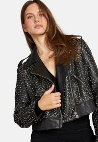 Giovane donna con lunghi capelli castani che indossa una giacca di pelle nera con borchie sopra un top nero, in posa su uno sfondo bianco.