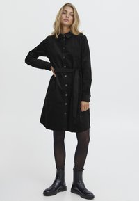 Robe chemise en velours côtelé noir avec un front à boutons, des manches longues et une ceinture assortie. Finition avec un ourlet arrondi et des poches.