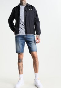Svart lättviktsjacka över en grå t-shirt med uggletryck, tillsammans med blå denimshorts och vita sneakers. Tatueringar synliga på armarna.