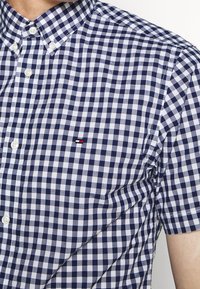 Hombre vistiendo una camisa de botones de manga corta a cuadros en azul marino y blanco, con un pequeño logo rojo, blanco y negro en el pecho.