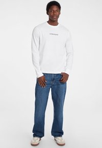 Hvid langærmet skjorte med "GUESS JEANS" trykt på brystet, parret med løstsiddende blå denim jeans og brune sneakers.